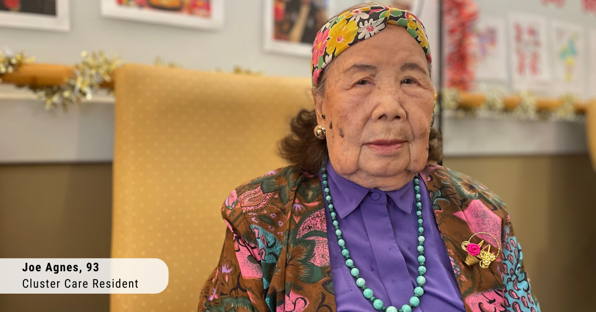 an older asian woman wearing a flowered head scarf, flowered blazer and purple blouse and blue beads sits facing the camera