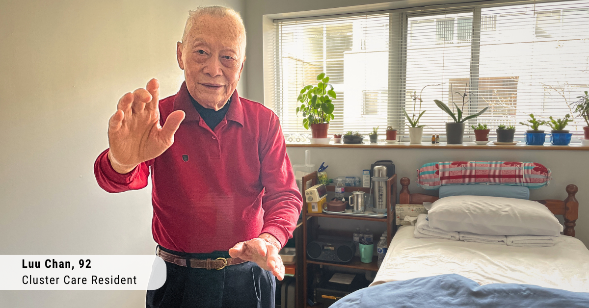 an older asian man wearing a red shirt stands in front of a bed with a window behind it and is doing tai chi moves