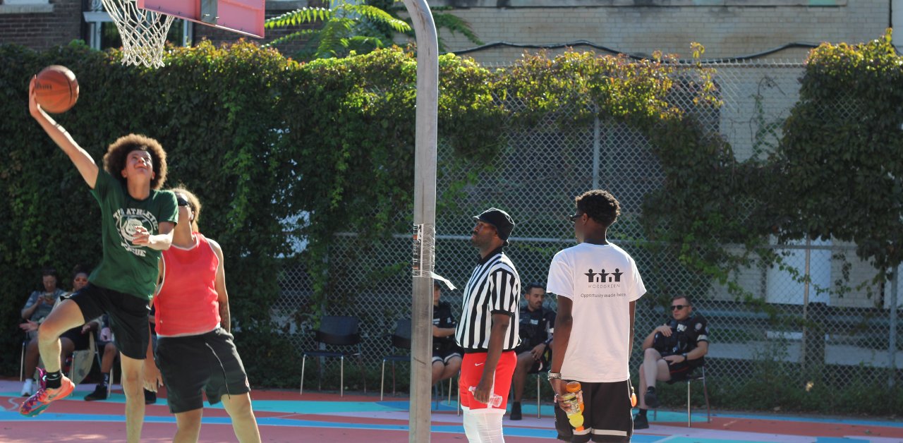 A teenaged boy jumps with a basketball on a court as a referee and two other players look on, with Toronto police officers sitting on the sidelines as spectators.