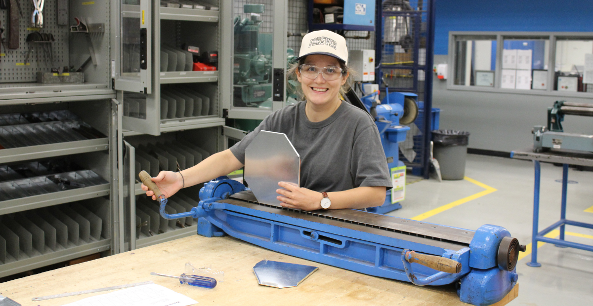 a woman in a grey shirt wearing a cap and safety goggles works on a sheet metal press during a plumbing program at WoodGreen and George Brown College